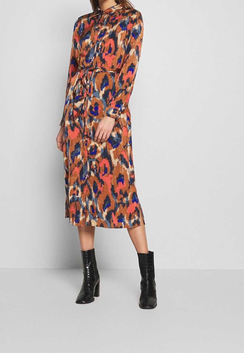 Femme portant une robe midi à manches longues boutonnée avec un motif abstrait orange, bleu et marron, associée à des bottines noires à talons.
