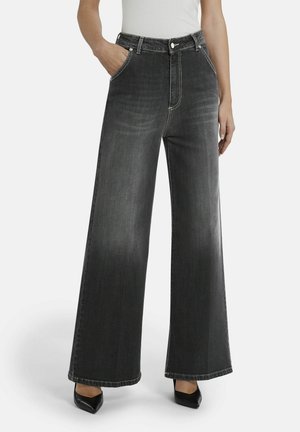 Frau trägt hoch taillierte, weit geschnittene schwarze Jeans mit ausgewaschenen Details, weißen, eingesteckten Oberteil, Hände in den Taschen und schwarze spitz zulaufende Schuhe.