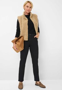 Gilet en fausse fourrure beige sur un pull noir, assorti à un jean noir et des chaussures à imprimé léopard. Portant un sac en daim marron.