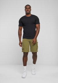 Un model care stă poartă un tricou scurt negru cu mâneci scurte, pantaloni scurți cargo de culoare verde măsliniu cu buzunare laterale și adidași albi cu tălpi texturate.