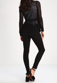 Musta avoin neulepaita pitkät hihat, jossa on teksturoitu kuvio, kerrottu mustien tarkasti istuvien housujen päällä ja mustien korkeakorkoisten kenkien, joissa on nauhaaksentteja.