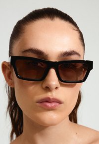 Ochelari de soare dreptunghiulari negri, cu rame groase și lentile maro, având un design elegant și colțuri unghiulare. Fără embellishments suplimentare.