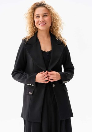 Black blazer with a wide collar, structured design, and silver buttons. Textured fabric, featuring a pocket with a brand label.