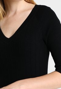 Haut en tricot noir à côtes avec un décolleté en V et des manches courtes. La texture est lisse, avec des lignes verticales sur l'ensemble du tissu.