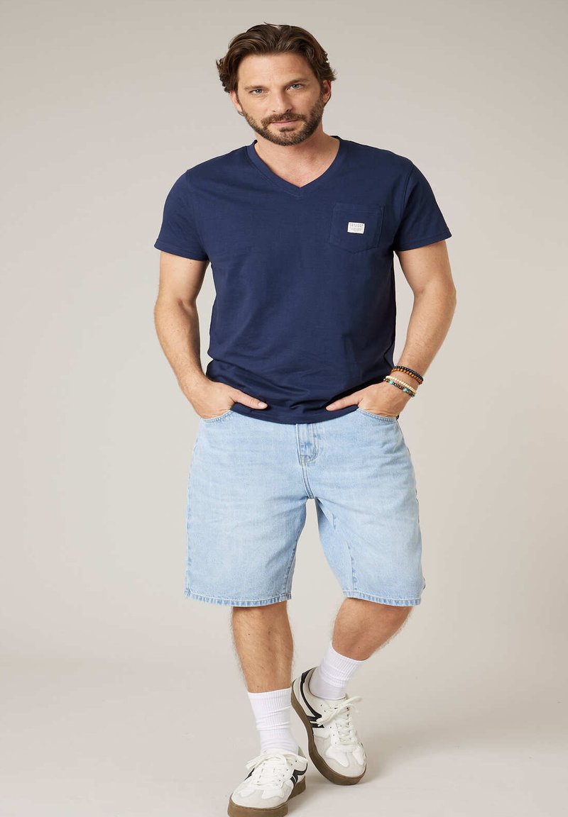 Homme portant un t-shirt bleu marine à col en V, un short en denim bleu clair, des baskets blanches et des bracelets en perles, debout les mains dans les poches.