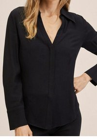 Blouse noire à manches longues en tissu lisse. Présente un col en V, un col classique et des poignets avec boutons. Coupe droite avec de petites fentes sur les côtés.