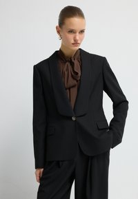 Zwarte blazer met een piek-lapels, streeppatroon, enkele knoopsluiting en twee voorzakken, gedragen over een bruine blouse.