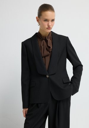 Blazer noir avec un revers pointu, motif à rayures, fermeture à un bouton, et deux poches avant, porté sur un chemisier marron.