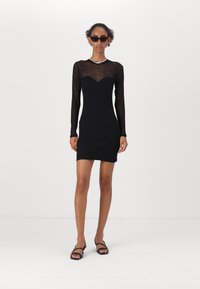 Black dress with long sheer sleeves, sweetheart neckline, and embellishments. Fitted design, smooth texture, paired with black sandals.
