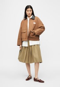 Γυναίκα που φοράει υπερμεγέθη καφέ μπουφάν με μαύρο γιακά, άσπρο πουκάμισο, πλισέ καφέ φούστα και σκουρόχρωμα καφέ παπούτσια Mary Jane, στέκεται σε λευκό φόντο.