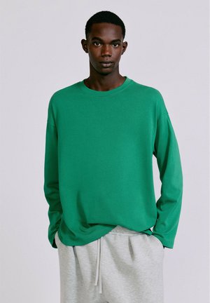 Jeune homme portant une chemise verte ample à manches longues et un pantalon de survêtement gris clair, debout avec les mains dans les poches.