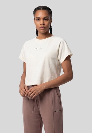 Vrouw met gevlochten haar die een wit cropped Champion T-shirt en mauve Champion joggingbroek draagt tegen een grijze achtergrond.
