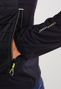 Sort jakke med lynlåslommer, lavet af letvægtsmateriale og med struktureret ærme. Logoet i gult er vist på ærmet.