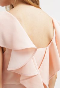 Svetle růžové šaty s vrstveným volánkovým designem, s V- výstřihem na zádech a jemnou, texturovanou tkaninou. Detailní záběr ukazuje splývavou siluetu oděvu.