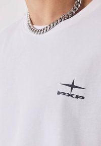 Camiseta blanca de algodón con un pequeño logotipo negro "PXP" y un diseño de estrella en el lado izquierdo del pecho, combinada con un collar de cadena de plata gruesa.