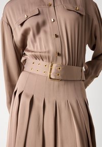 Beige pleated dress, jossa on napitettava kaulus ja kultaiset yksityiskohdat. Mekkoon kuuluu yhteensopiva vyö, jossa on pyöreitä reikiä ja suorakulmainen solki.