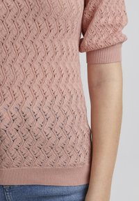 Roze gebreide trui met een golfpatroon, voorzien van korte mouwen en een geribbelde zoom. De textuur is zacht en licht doorschijnend voor ademend vermogen.
