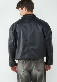 Giacca nera in pelle sintetica con colletto, vestibilità rilassata e dettagli di cucitura in evidenza. Abbinata a pantaloni in denim grigio sbiadito.