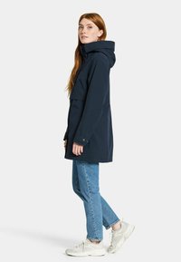 Marineblauwe waterdichte jas met kap, voorzien van drukknoopsluiting en borstzakken, gecombineerd met lichtblauwe spijkerbroek en beige sneakers.