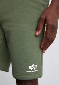Oliven grønne bomuldsbukser med ribbet tekstur, med hvid "Alpha Industries" logo og geometrisk emblem nær kanten. Hånd hviler på stoffet.