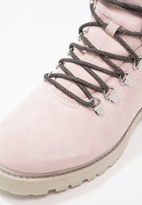 Bota de senderismo de ante rosa claro con cordones negros, ojales plateados y una suela gruesa de color crema con un patrón de relieve pronunciado.