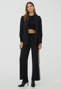 Donna che indossa un blazer nero, un maglione corto, pantaloni a gamba larga con striscia bianca laterale e stivaletti neri con tacco alto, in piedi su uno sfondo bianco.