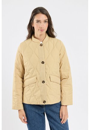 Femme portant une veste matelassée beige avec de gros boutons et des poches à rabat, associée à un jean bleu, debout devant un fond uni.