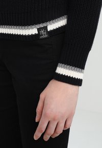 Pull en maille noire avec des poignets rayés blanc et gris. Présente un patch logo Superdry près de l'ourlet. Main reposant contre un pantalon noir.