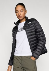 Μαύρο μπουφάν puffer με φερμουάρ μπροστά, με ραμμένες επιφάνειες και ψηλό γιακά. Συνδυάζεται με λευκό μπλουζάκι και χακί παντελόνι.