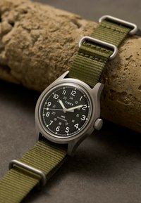 Reloj mecánico de pulsera Timex MK1 con esfera negra, números blancos, caja de plata y correa de tela verde, apoyado sobre una superficie beige rugosa.