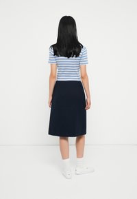 Žena s rovnými čiernymi vlasmi, oblečená v modro-bielom pruhovanom tričku s krátkym rukávom, námorníckej sukni po kolená, bielych ponožkách a bielych teniskách, otočená chrbtom.