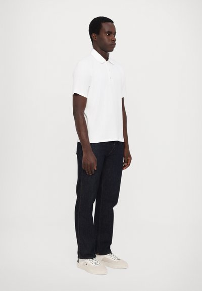 Polo shirt blanc à manches courtes, jeans bleu marine et baskets blanches. Design simple, tissu texturé et coupe standard.