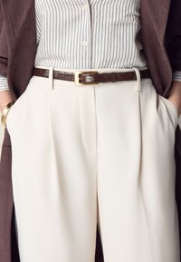 Vita plisserade byxor med ett brunt läderbälte med guldfärgade spänne, kombinerat med en randig skjorta och ett brunt ytterplagg. Slät tygstruktur.