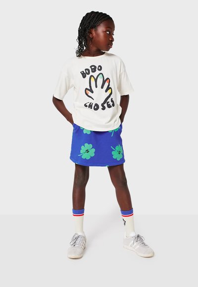 Vit t-shirt med svart handgrafik och färgglada fingrar, tillsammans med en blå kjol med gröna klövermönster. Grå sneakers och randiga strumpor.