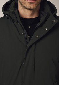 Mężczyzna w ciemnozielonej zimowej kurtce z kapturem, zapinanej na zamek błyskawiczny i napy, nosi czarną koszulę zapinaną na guziki. Zbliżenie na klatkę piersiową i podbródek.