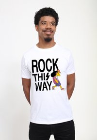 Muž v bílém tričku s nápisem „ROCK THIS WAY“ a kreslenou postavičkou s afrem, kloboukem, sluchátky a pohodlným oblečením, který kráčí.