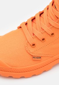 Las zapatillas de lona naranjas cuentan con un upper texturizado, cordones redondos y una suela de goma. Una pequeña etiqueta "PALLADIUM" es visible en el costado.
