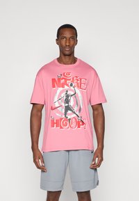 Ροζ βαμβακερή μπλούζα με γραφικό σχέδιο ενός μπασκετμπολίστα, με την επιγραφή "NIKE HOOPS" σε έντονες κόκκινες και μαύρες αποχρώσεις.