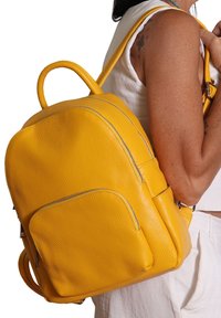 Femme à la peau medium portant un top sans manches blanc et un pantalon, portant un sac à dos en cuir jaune texturé sur une épaule.