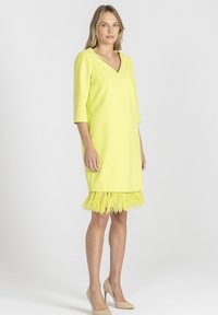 Kobieta w żółtej sukience do kolan z rękawami o długości trzech ćwiartek oraz wykończeniem z piór, w połączeniu z beżowymi szpilkami, stoi na tle białym.