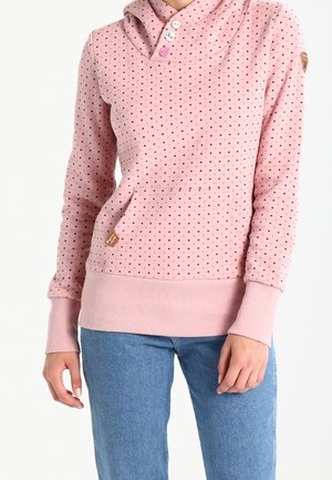 Femme portant un sweat à capuche rose à pois avec des boutons et un jean bleu, debout les mains détendues sur les côtés.