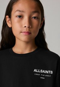 Juodas medvilninis marškinėliai su balta spauda: "ALLSAINTS," su japonų simboliais ir "LONDON" apačioje. Klasikinio apvalaus kaklo dizainas.