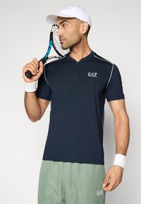 Μπλε ναυτικό αμάνικο αθλητικό πουκάμισο με φερμουάρ μπροστά και λευκές λεπτομέρειες, συνδυασμένο με σορτς σε μέντα πράσινο χρώμα. Ο παίκτης κρατά ρακέτα του τένις.