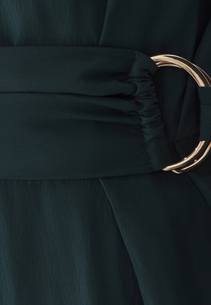 Dunkelgrünes Kleid mit einem gerafften Bund, der mit einer großen goldenen Metallschnalle verziert ist. Der Stoff wirkt leicht und hat eine glatte Textur.