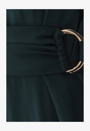 Abito verde scuro con una vita arricciata e una grande fibbia in metallo dorato. Il tessuto sembra leggero e ha una superficie liscia.