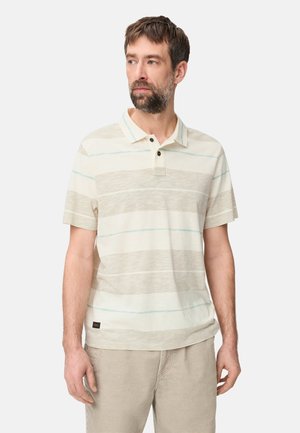 Mann, der ein kurzärmeliges Polohemd mit beigen und weißen Streifen und beige Hose trägt, schaut leicht nach links vor einem schlichten Hintergrund.