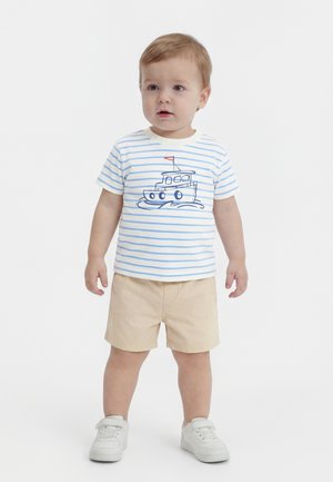 Kleinkind steht in beigen Shorts, weißen Turnschuhen und einem weiß-blau gestreiften T-Shirt mit einem Boot-Motiv.