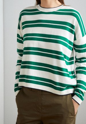 Personne portant un pull blanc à rayures horizontales vert foncé et un pantalon marron, les mains dans les poches, debout contre un mur uni.