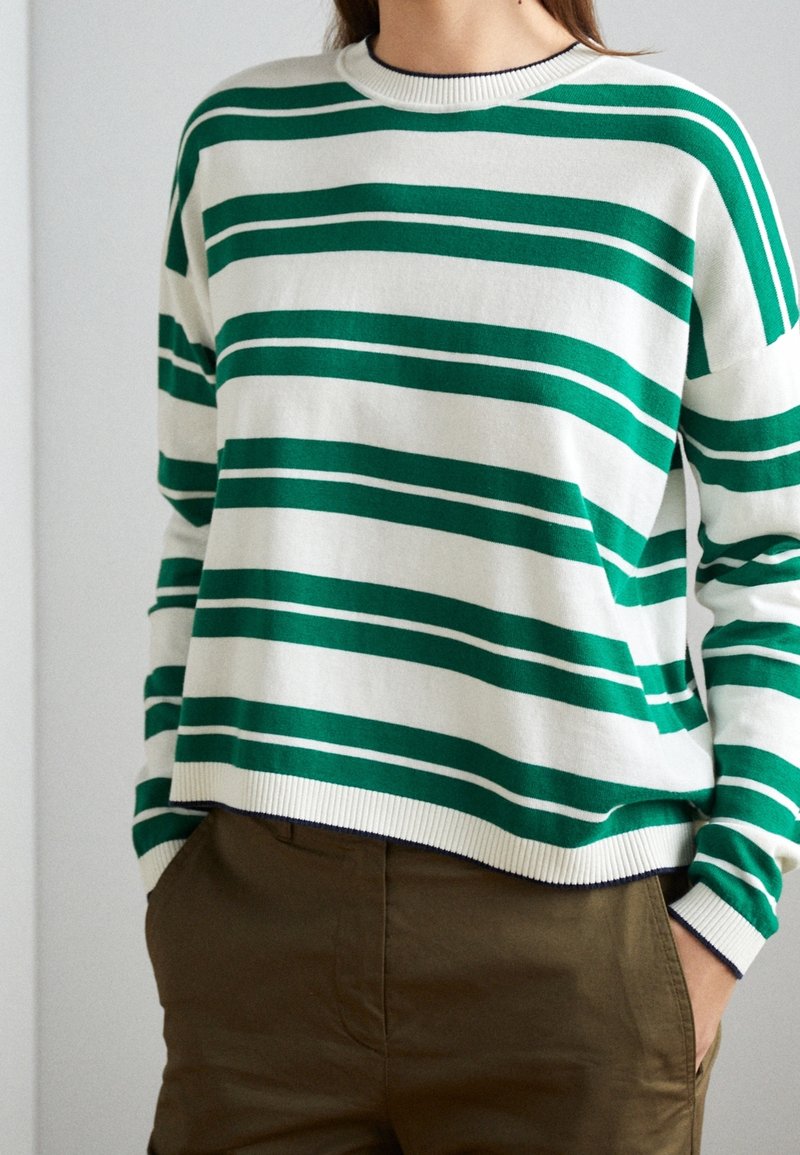 Personne portant un pull blanc à rayures horizontales vert foncé et un pantalon marron, les mains dans les poches, debout contre un mur uni.