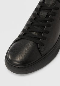 Μαύρο αθλητικό παπούτσι από δέρμα με υφή στην επιφάνεια, μαύρα κορδόνια και χοντρή λαστιχένια σόλα σε απλό ανοιχτό φόντο.